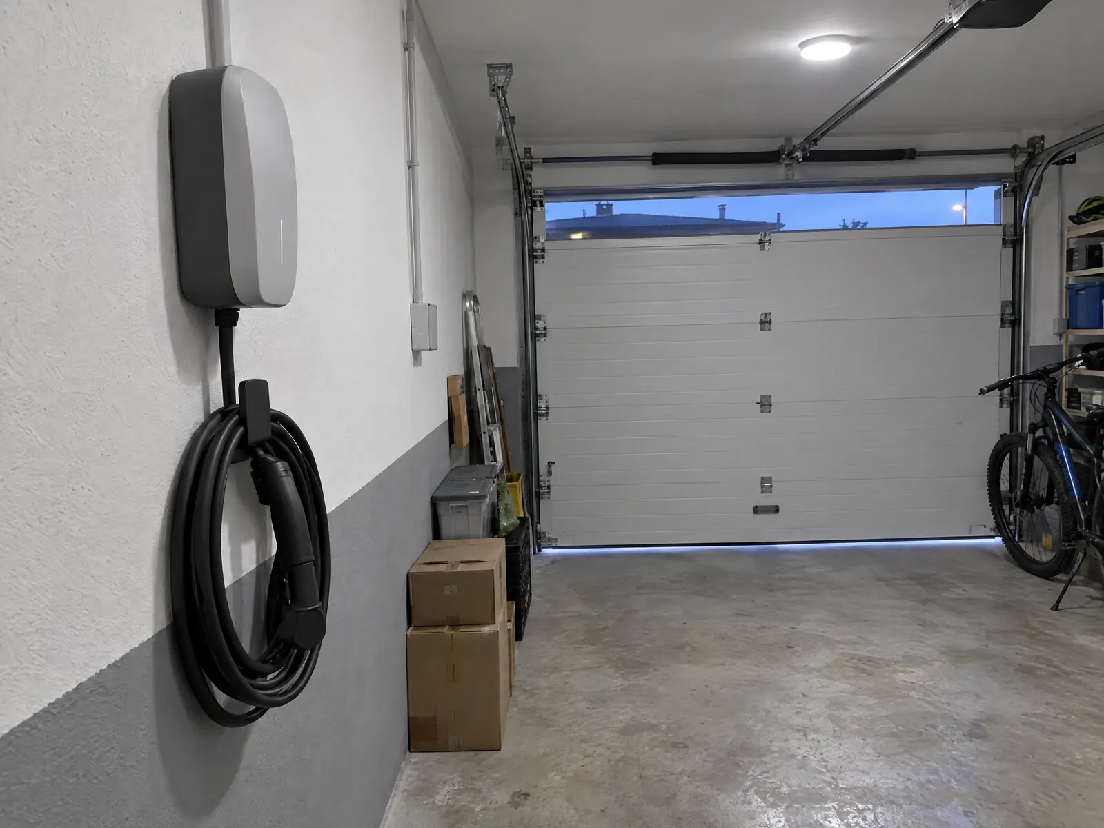 Garaje de una vivienda con un cargador de coche eléctrico en la pared y su manguera enrollada, puerta seccional al fondo