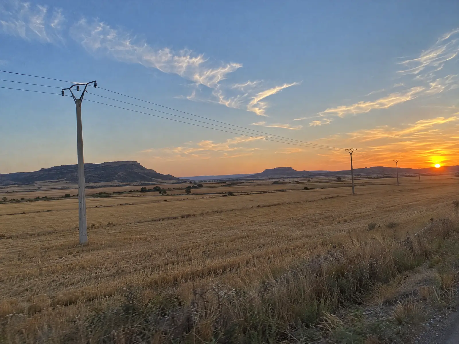 Campos de cereal segados al atardecer con un tendido eléctrico sobre postes y cerros planos en el horizonte