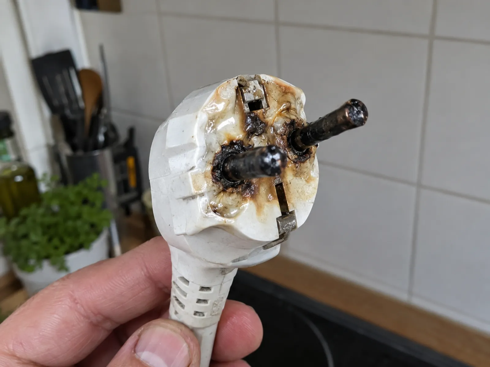 Mano sujetando una clavija blanca con las patillas ennegrecidas y el plástico fundido y marrón alrededor, en una cocina