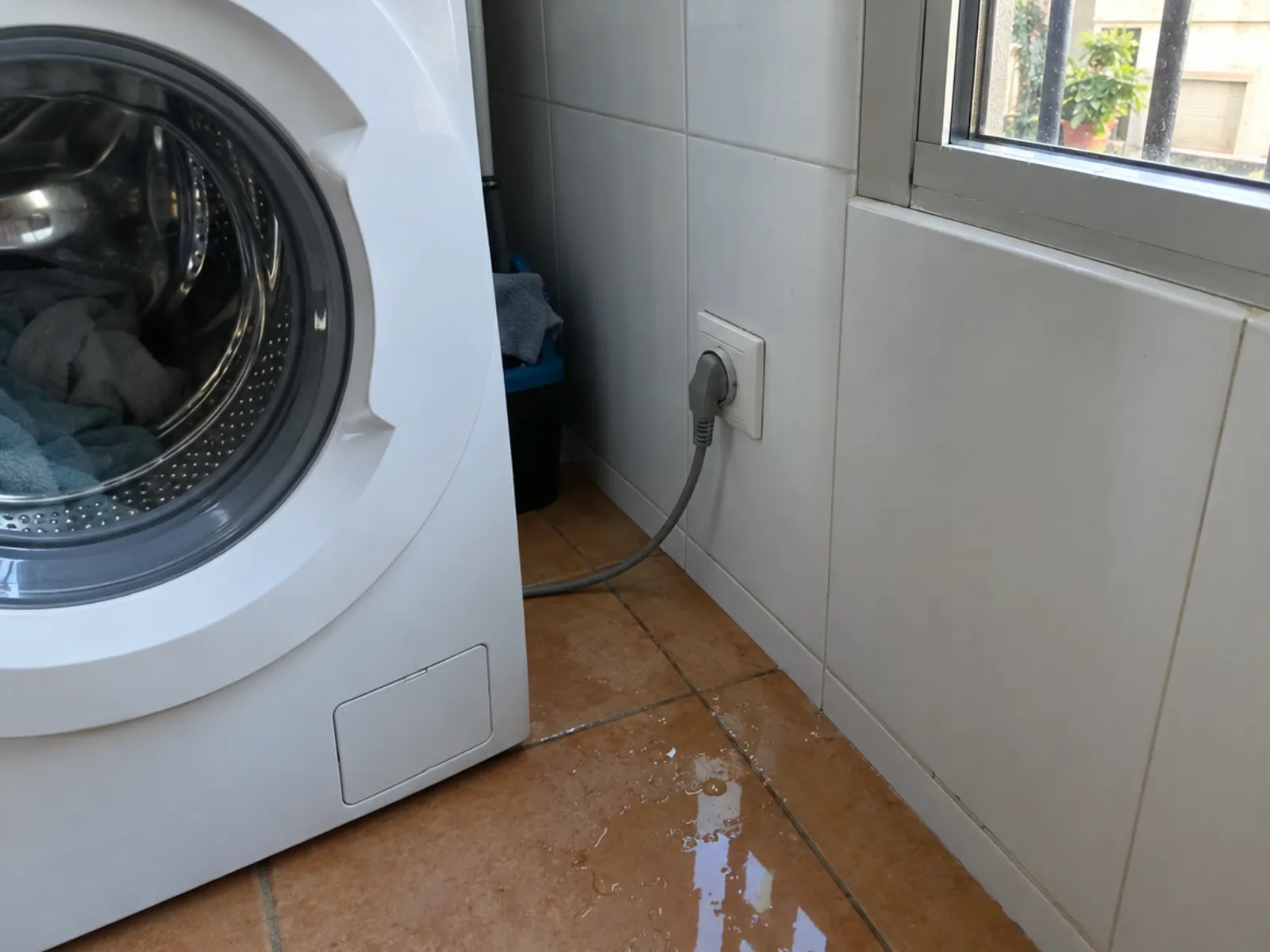 Lavadora en un lavadero alicatado enchufada a una toma de pared junto al suelo, con un charco de agua en las baldosas