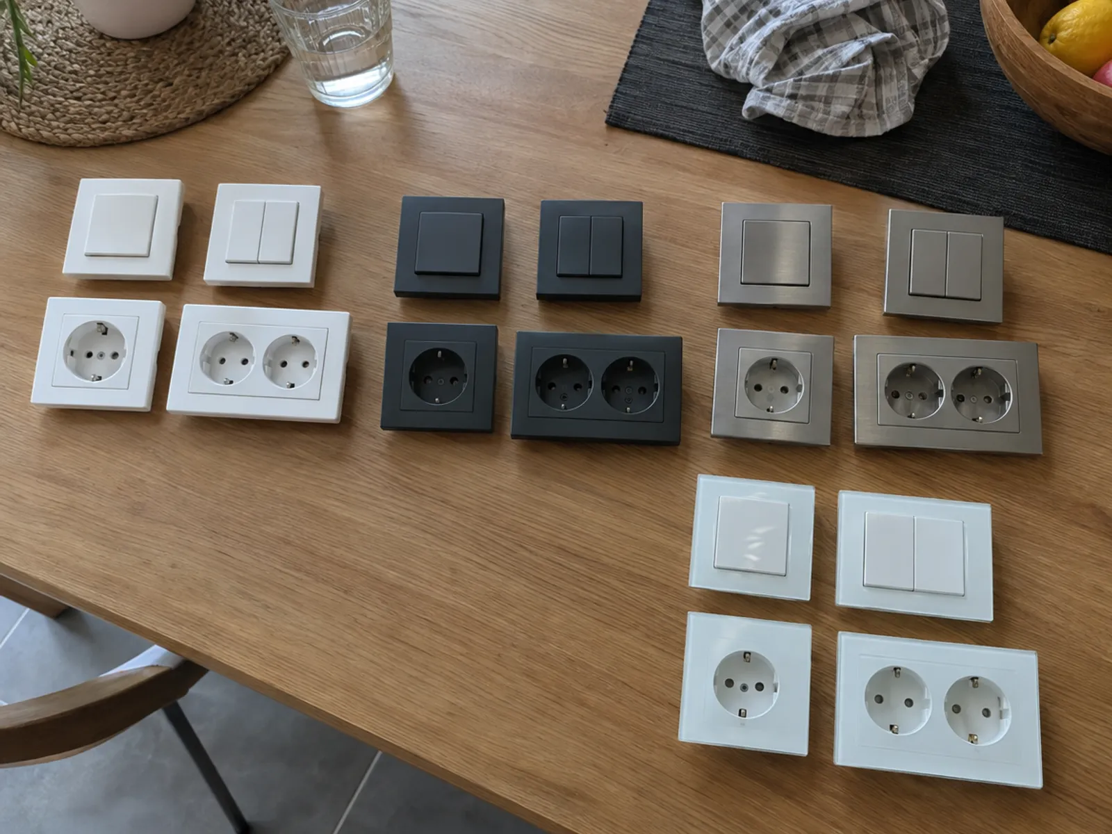 Interruptores y enchufes de cuatro acabados distintos colocados sobre una mesa de madera: blanco, antracita, acero y cristal blanco