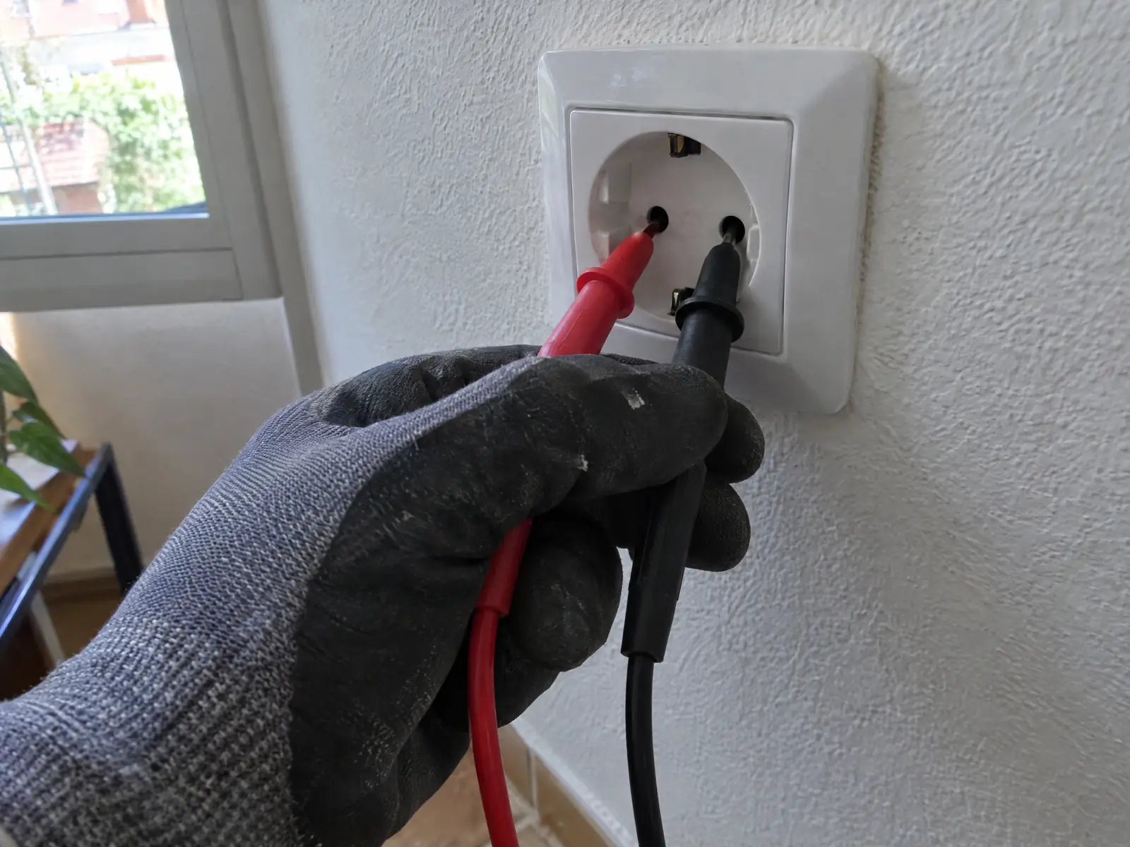 Mano con guante introduce las puntas roja y negra de un polímetro en los alveolos de un enchufe de pared