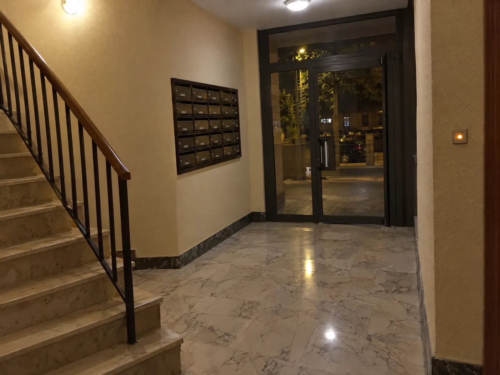 Portal de un edificio de noche con la luz encendida, buzones, escalera de mármol y el pulsador de la luz iluminado