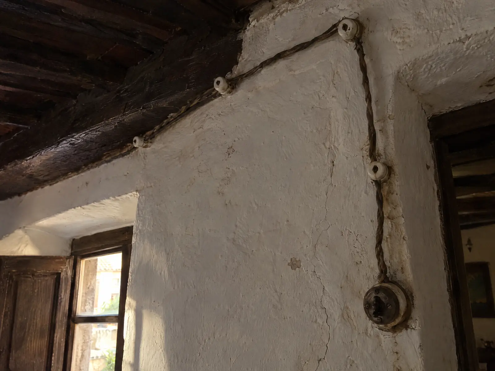 Muro encalado con viga de madera y cable trenzado antiguo grapado sobre aisladores de porcelana hasta un interruptor de superficie